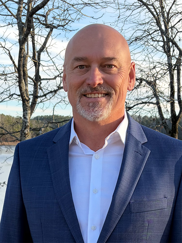 Professional outdoor headshot showing Troy Chastain wearing a blue suit jacket and white button‑down shirt, standing in front of bare trees and a lake.