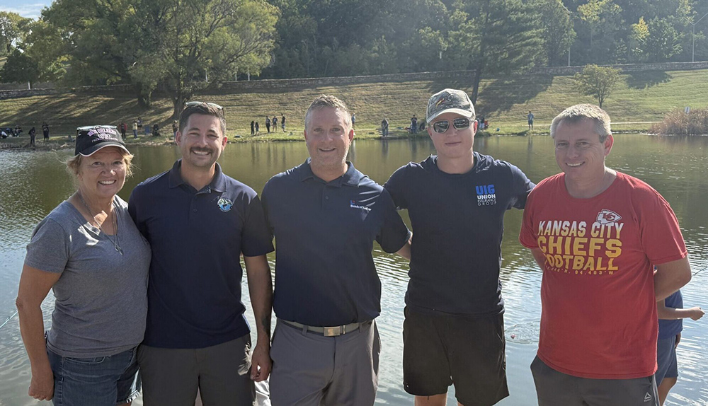 Group of 5 people smiling in front of a lake. Johnson (third from left) with other volunteers at the 2025 Greater Kansas City BCTC Take Kids Fishing Day.